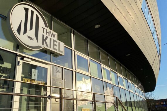 Das Bild zeigt die moderne Glas- und Metallfassade der THW-Sportarena Kiel, mit dem Vereinslogo und dem Schriftzug THW KIEL 1904 über dem Eingang.