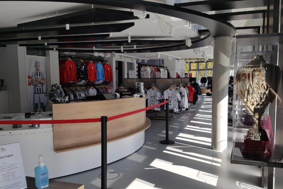 Ein modernes Sportartikelgeschäft mit Trikots und Kleidung in Regalen, einer Theke mit Handdesinfektionsmitteln und einem großen Pokal in einer Glasvitrine neben dem Eingang. Durch große Fenster strömt Sonnenlicht.