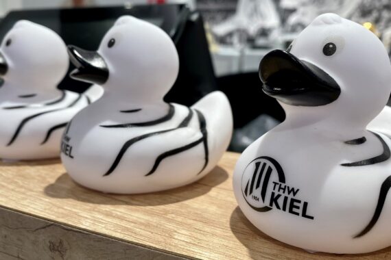 Drei weiße Gummi-Enten mit schwarzen Schnäbeln und Streifen sind auf einer Holzfläche aufgereiht. Die nächstgelegene Ente hat ein THW KIEL-Logo auf der Seite. Der Hintergrund ist unscharf.
