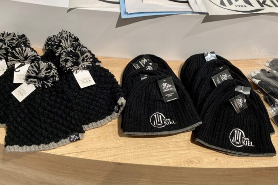 Gestrickte schwarze und graue Bommelmützen und einfarbige schwarze Mützen mit THW Kiel Logo auf einer Holzfläche, rechts ein Paar schwarze Handschuhe.