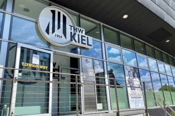 Außenansicht des THW Kiel-Gebäudes mit Glasfenstern, einem großen runden THW Kiel 1904-Logo über dem Eingang, Geländer und einem Plakat mit Menschen auf der rechten Seite. Der blaue Himmel mit Wolken spiegelt sich im Glas.