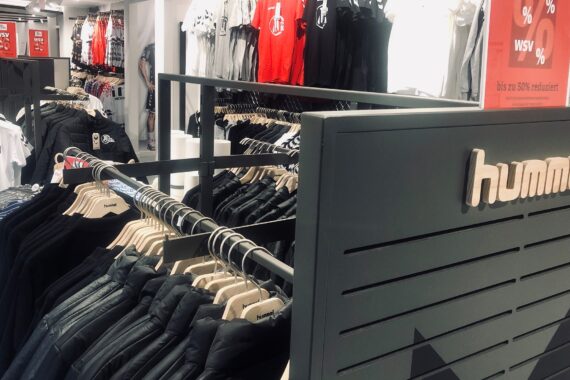Innenraum eines Bekleidungsgeschäfts mit Regalen mit Jacken und Hemden. Die Marke hummel ist auf einem Schild abgebildet. Rote Verkaufsschilder in deutscher Sprache weisen auf Rabatte von bis zu 50 % hin. Die Kleidungsstücke sind fein säuberlich auf Bügeln angeordnet.