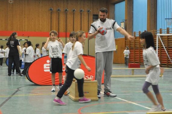 Kinder und ein erwachsener Trainer nehmen an einer sportlichen Aktivität in einer Turnhalle teil. Kinder in weißen Hemden spielen mit Bällen und Holzklötzen, während andere im Hintergrund zuschauen oder mitmachen.