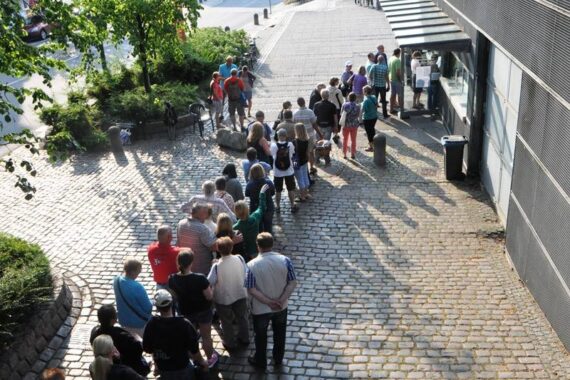 Eine lange Schlange von Menschen steht auf einem Kopfsteinpflasterweg vor einem Gebäude mit großen Fenstern und wartet darauf, einzutreten oder etwas zu kaufen. Auf der linken Seite des Weges sind Bäume und Grünflächen zu sehen.