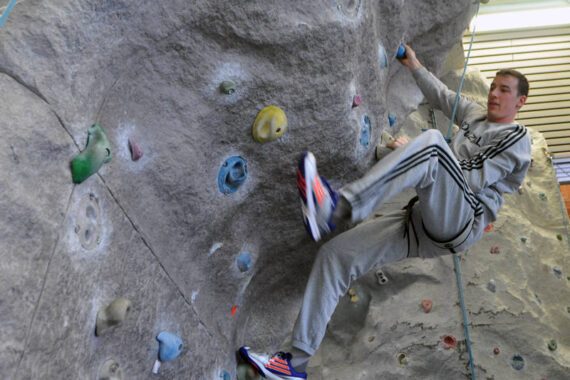 Eine Person, die einen grauen Trainingsanzug und bunte Turnschuhe trägt, klettert an einer Indoor-Felswand, wobei sie sich mit einer Hand und einem Fuß an Griffen festhält und das andere Bein nach außen streckt. Die Kletterwand hat verschiedenfarbige Griffe.