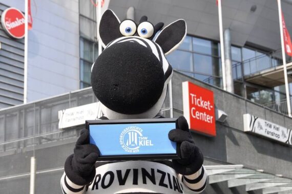 Eine Person in einem Zebra-Maskottchen-Kostüm hält ein blaues Schild mit dem Logo des THW Kiel vor einem Gebäude mit einem Ticket-Center-Schild und mehreren roten und weißen Fahnen.