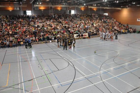 Eine überfüllte Sporthalle mit vielen Zuschauern auf den Tribünen, die während eines Spiels oder einer Veranstaltung zwei Mannschaften auf einem Basketballfeld beobachten.