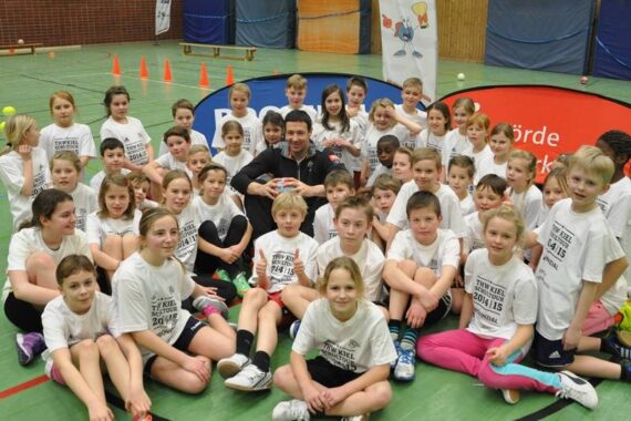 Eine Gruppe von Kindern in passenden weißen T-Shirts sitzt und kniet auf dem Boden einer Turnhalle und lächelt in die Kamera. Ein Erwachsener sitzt in der Mitte, und im Hintergrund sind Sportgeräte zu sehen.