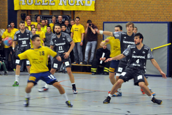 Ein Handballspiel ist im Gange; ein Spieler in Gelb bereitet sich auf den Wurf vor, während vier Spieler in Grau den Ball verteidigen. Die Zuschauer schauen von der Tribüne aus zu, im Hintergrund ist ein gelbes Banner mit der Aufschrift Hölle Nord zu sehen.