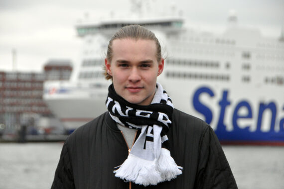Eine junge Person mit langen, hellen Haaren, die nach hinten gebunden sind und eine schwarze Jacke und einen schwarz-weißen Schal tragen, steht im Freien vor einer großen weißen Fähre mit der Aufschrift Stena.