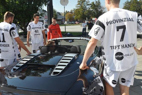 Eine Gruppe männlicher Athleten in weißen Sportuniformen steht um ein schwarzes Cabrio herum, unterhält sich und lächelt an einem sonnigen Tag mit Bäumen und Gebäuden im Hintergrund.