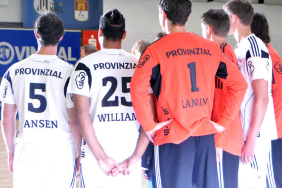 Eine Gruppe männlicher Sportler, die weiße und orangefarbene Trikots mit Nummern und dem Wort "Provinzial" auf dem Rücken tragen, stehen in einer hell erleuchteten Turn- oder Sporthalle zusammen und schauen von der Kamera weg.