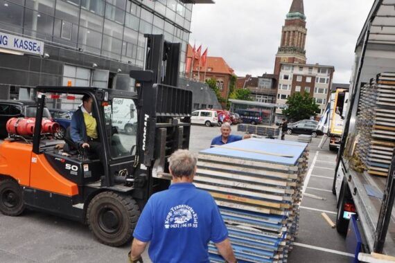 Arbeiter entladen mit einem Gabelstapler große Metall- und Holzplatten von einem Lkw auf einem Parkplatz neben einem modernen Gebäude und einem historischen Uhrenturm im Hintergrund.