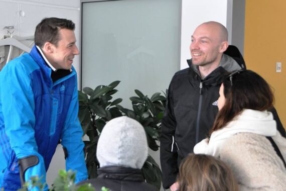 Vier Erwachsene in Winterkleidung stehen in einem Haus, lächeln und unterhalten sich. Zwei Kinder mit Mützen und Mänteln stehen im Vordergrund und wenden sich ab. Im Hintergrund sind Grünpflanzen und eine gelbe Wand zu sehen.