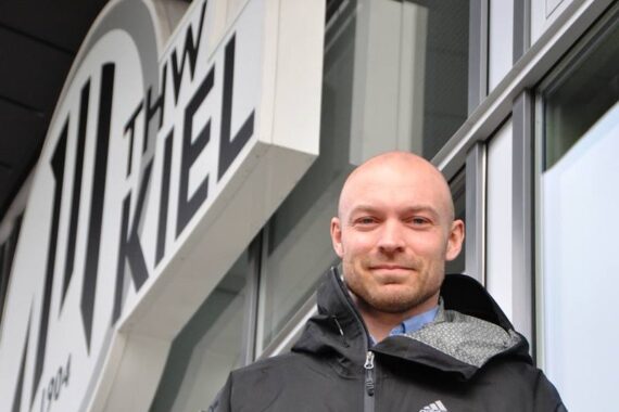 Ein Mann in einer schwarzen Adidas-Jacke steht vor einem Gebäude mit einem großen THW Kiel-Schild. Er hat eine Glatze und lächelt leicht, und im Hintergrund sind Glasfenster und ein Teil des Schildes zu sehen.