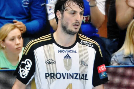 Ein männlicher Handballspieler mit nassen Haaren, der ein schwarz-weißes Trikot mit der Nummer 4 und dem Wort PROVINZIAL auf der Vorderseite trägt, steht auf dem Spielfeld. Im unscharfen Hintergrund sind Zuschauer zu sehen.