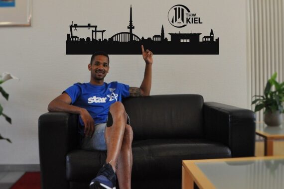 Ein Mann im blauen T-Shirt sitzt entspannt auf einem schwarzen Sofa und zeigt auf ein Wandtattoo mit dem Logo des THW Kiel und einer Stadtsilhouette. In dem Raum sind Zimmerpflanzen und ein kleiner Tisch zu sehen.