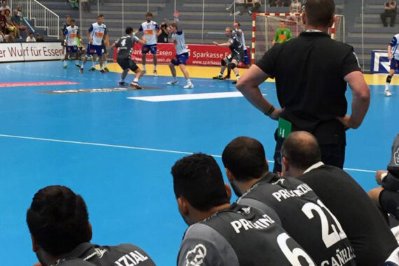 Handballspiel der Männer auf einem blauen Spielfeld. Spieler in weißen und blauen Trikots spielen, während Spieler in schwarzen Trikots mit der Aufschrift "PRONZIAL" auf dem Rücken von der Bank aus zusehen. Ein Trainer steht in der Nähe.
