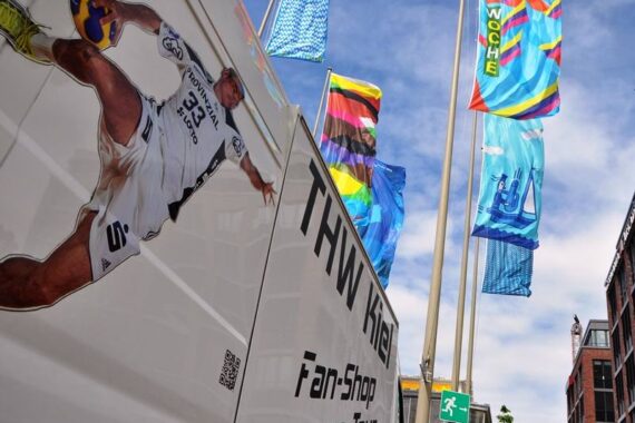 Ein Lieferwagen mit einem großen Bild eines Handballspielers und dem Schriftzug THW Kiel Fan-Shop on Tour parkt neben bunten Fahnen, die im Wind wehen, im Hintergrund sind Gebäudeteile zu sehen.