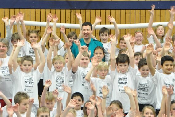 Ein lächelnder Erwachsener steht inmitten einer großen Gruppe von Kindern, die alle passende weiße Hemden tragen und ihre Hände in die Luft heben, in einem Raum mit einer Holzwand und einem Torpfosten im Hintergrund.