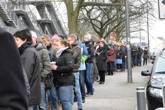 Eine lange Schlange von Menschen in Winterkleidung wartet an einem bewölkten Tag auf einem Bürgersteig in der Nähe geparkter Autos und blattloser Bäume.