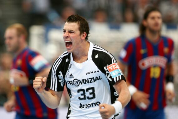 Ein männlicher Handballspieler in einem weiß-schwarzen Trikot mit der Nummer 33 ballt die Fäuste und jubelt auf dem Spielfeld, während im Hintergrund gegnerische Spieler in blauen und roten Trikots zu sehen sind.