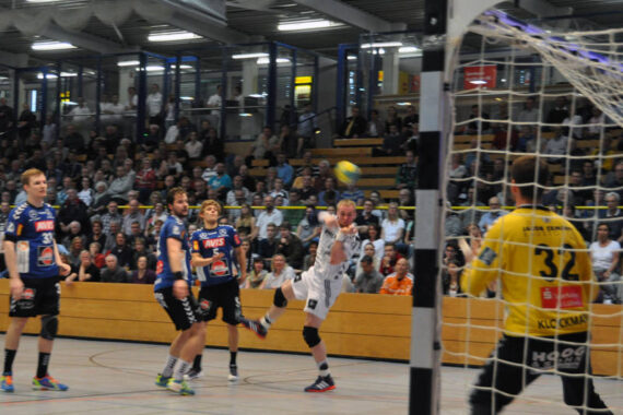 Ein Handballspieler in einem weißen Trikot springt, um den Ball in Richtung Tor zu schießen, während gegnerische Spieler in blauen Trikots zusehen und der Torwart in einem gelben Trikot sich darauf vorbereitet, den Ball abzublocken, während die Zuschauer im Hintergrund sitzen.