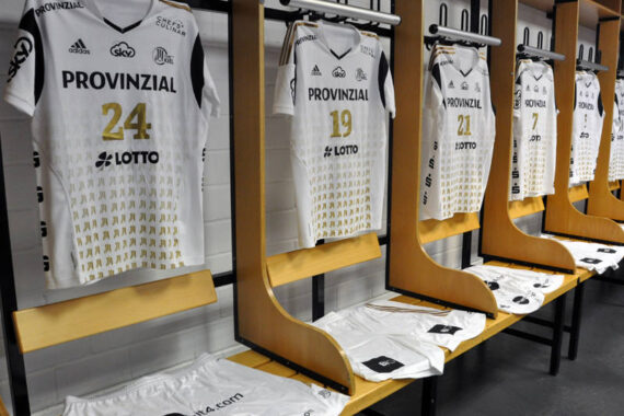 In einer Umkleidekabine hängen mehrere weiße Sporttrikots mit Nummern und Sponsorenlogos in einer Reihe, unter denen auf den Holzbänken passende Shorts liegen.