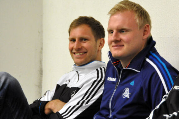 Zwei Männer sitzen nebeneinander vor einer weißen Wand, beide tragen Sportjacken und lächeln. Der Mann auf der linken Seite hat hellbraunes Haar und eine Jacke mit Reißverschluss, der Mann auf der rechten Seite hat blondes Haar und eine blaue Jacke mit Logos.