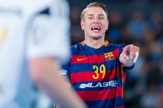 Ein Handballspieler in einem blau-rot gestreiften Trikot mit der Nummer 39 zeigt während eines Spiels auf einen anderen Spieler in einem weißen Trikot, der im Vordergrund steht, und schreit.