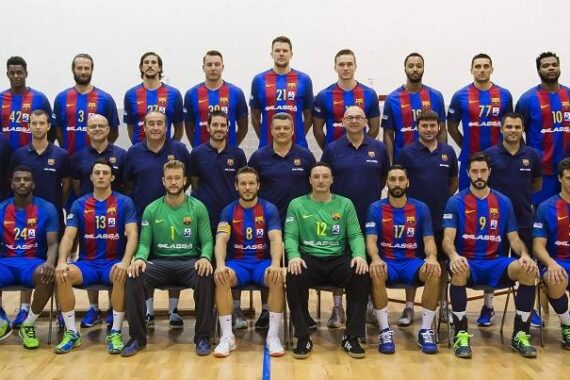 Eine Herren-Handballmannschaft in blauen und roten Trikots posiert mit ihren Trainern auf einem Holzplatz. Die Gruppe ist in drei Reihen angeordnet, wobei die Spieler und Trainer sitzen und stehen.