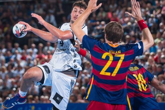 Ein Handballspieler in einem weißen Trikot springt, um den Ball in Richtung Tor zu werfen, während zwei Verteidiger in rot und blau gestreiften Trikots ihre Arme heben, um ihn während eines überfüllten Spiels zu blockieren.