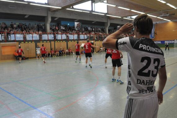 Ein Handballspieler im weißen Trikot mit der Nummer 23 steht auf dem Spielfeld und deutet mit der Hand auf seinen Kopf, während andere Spieler in Rot und Weiß spielen und die Zuschauer von der Tribüne aus zusehen.