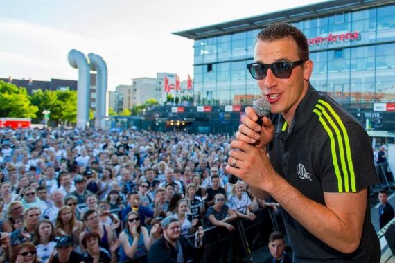 Ein Mann, der eine Sonnenbrille und ein schwarzes Hemd mit Neonstreifen trägt, spricht in ein Mikrofon auf der Bühne vor einer großen, jubelnden Menge vor einem modernen Gebäude mit der Aufschrift Arena.