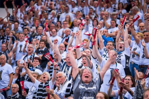 Eine große Menge begeisterter Sportfans, viele in passenden Trikots und Hemden, jubeln mit erhobenen Armen und schwenken rote Klatschen in einem Stadion. Die Szene ist voller Energie und Aufregung.