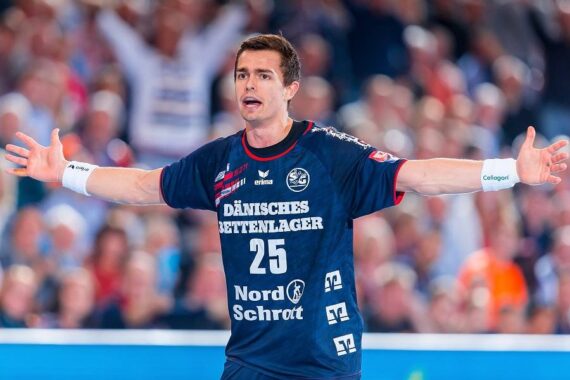 Ein männlicher Handballspieler in einem marineblauen Trikot mit der Nummer 25 hebt seine Arme weit, als wolle er während eines Spiels kommunizieren oder reagieren. Der Hintergrund zeigt eine unscharfe Menge und eine Halle.