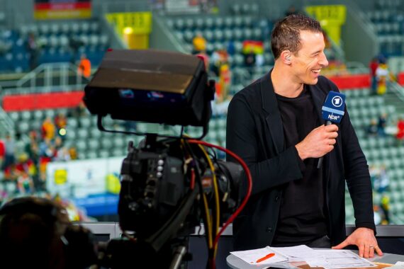 Ein lächelnder TV-Sportmoderator hält ein Mikrofon und Papiere in der Hand, während er am Set spricht, mit einer Kamera im Vordergrund und unscharfen Stadionsitzen und Zuschauern im Hintergrund.