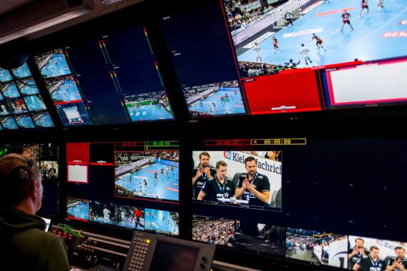 Eine Person bedient ein Schaltpult in einem Fernsehstudio, das mit Bildschirmen gefüllt ist, auf denen Live-Aufnahmen und mehrere Kameraperspektiven eines Handballspiels zu sehen sind und die Kommentatoren sprechen.