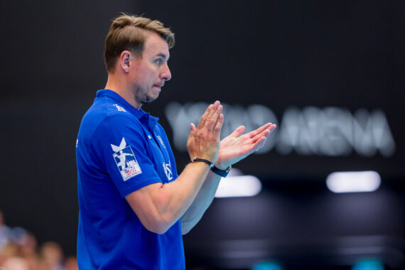 Ein Mann in einem blauen Poloshirt klatscht in die Hände und wirkt konzentriert. Er steht in einem Innenraum vor einem unscharfen Hintergrund, und hinter ihm ist der Teiltext XARD ARENA zu sehen.