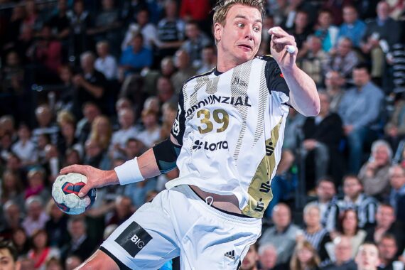 Ein männlicher Handballspieler in einem weiß-schwarzen Trikot springt in die Luft, hält einen Handball und will ihn werfen. Im Hintergrund ist eine unscharfe Zuschauermenge zu sehen, die das Spiel verfolgt.