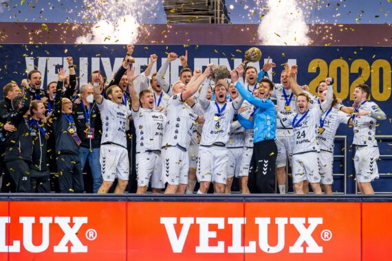 Eine Handballmannschaft der Männer feiert auf einem Podium und hebt eine Trophäe inmitten von Konfetti und Feuerwerk. Sie tragen weiße Trikots und Medaillen mit dem Schriftzug "VELUX" und "2020" im Hintergrund.