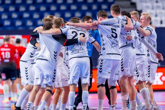 Eine Gruppe von männlichen Handballspielern in weißen Trikots liegt sich auf einem Hallenplatz in den Armen und jubelt. Im Hintergrund sind leere Tribünen und ein paar gegnerische Spieler in roten Trikots zu sehen.