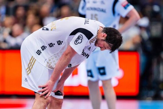 Ein männlicher Handballspieler in einem weißen Trikot beugt sich nach vorne und stützt die Hände auf die Knie. Er wirkt erschöpft oder enttäuscht während eines Spiels. Ein anderer Spieler steht im unscharfen Hintergrund.