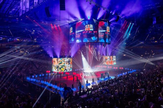 Eine große überdachte Arena mit einem jubelnden Publikum, bunten Bühnenlichtern und großen Bildschirmen. Die Darsteller stehen unter hellen Scheinwerfern auf der Bühne, Lichtstrahlen und leuchtende Farben erhellen die Szene.