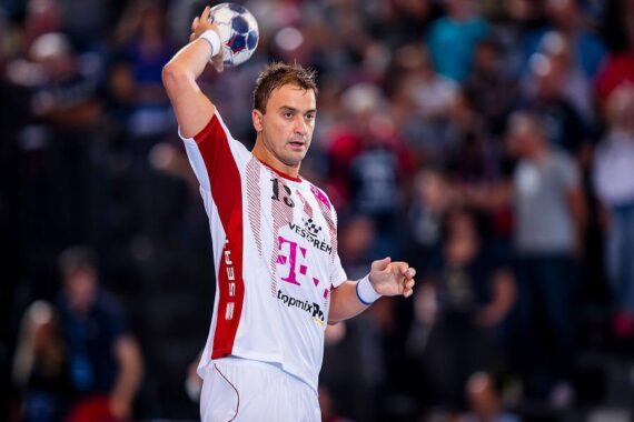 Ein männlicher Handballspieler in einem weiß-roten Trikot bereitet sich darauf vor, den Ball während eines Spiels zu werfen, während das Publikum im Hintergrund verschwommen zu sehen ist.