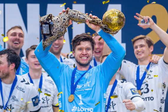 Ein lächelnder Handballspieler in einem blauen Trikot hebt eine große, goldene Trophäe in die Höhe, während seine Mannschaftskameraden in weißen Trikots um ihn herum feiern und goldenes Konfetti in die Luft fällt.