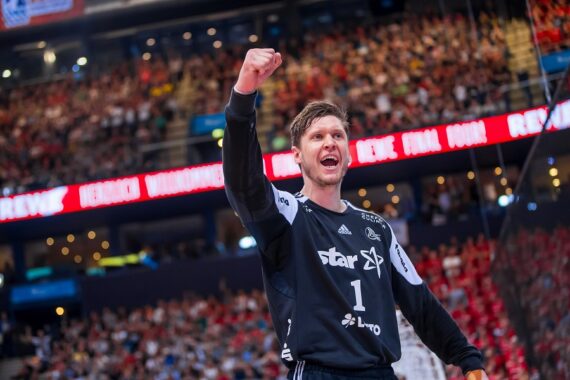 Ein männlicher Handballspieler in einem schwarzen Trikot jubelt mit erhobener Faust während eines Spiels in einer überfüllten Halle. Im Hintergrund sind die Zuschauer und leuchtende Fahnen zu sehen.