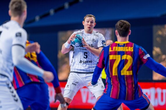 Ein Handballspieler in einem weißen Trikot hält den Ball, während er von zwei Spielern in blau-rot gestreiften Trikots bei einem Hallenspiel verteidigt wird. Der Fokus liegt auf dem Spieler, der sich auf einen Spielzug vorbereitet.