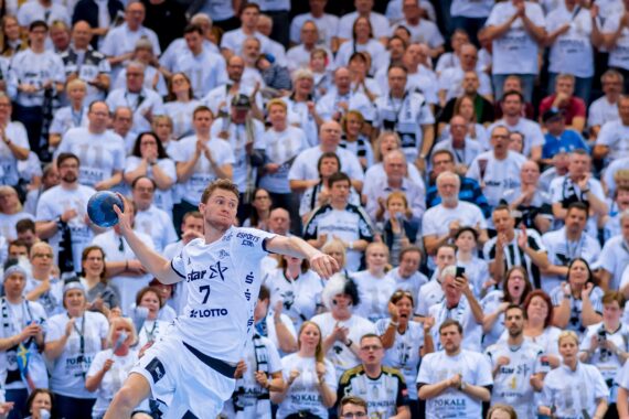 Ein Handballspieler in einem weißen Trikot springt in die Luft, um den Ball zu werfen, während im Hintergrund eine große Menge von Fans in passenden weißen Hemden jubelt.
