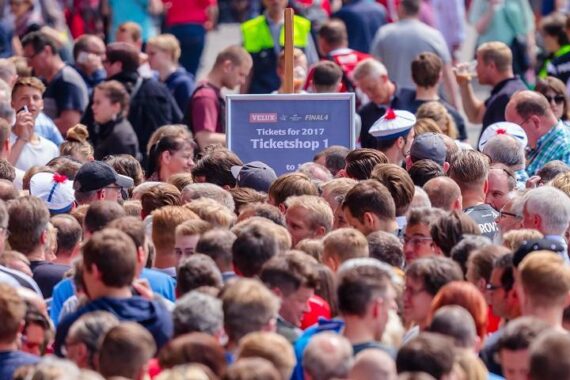 Eine große Menschenmenge steht dicht gedrängt im Freien, viele stehen vor einem Schild mit der Aufschrift "Tickets für 2017 - Ticketshop 1". Die Szene ist geschäftig und lebhaft, mit Menschen verschiedenen Alters.